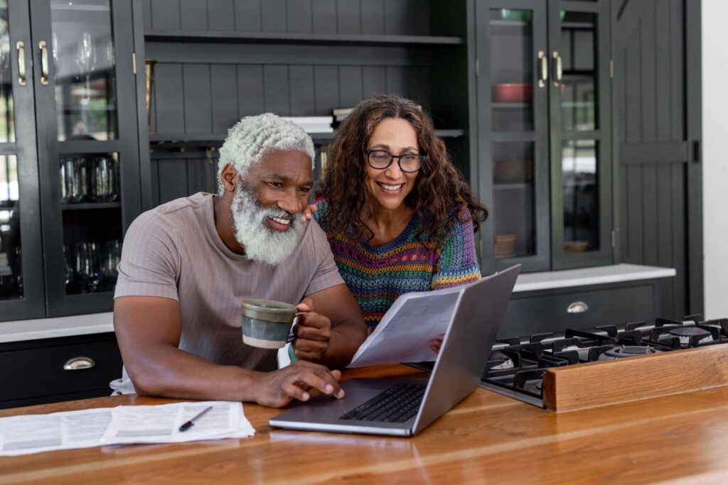 Older man and woman reading a book together at a table with a laptop.