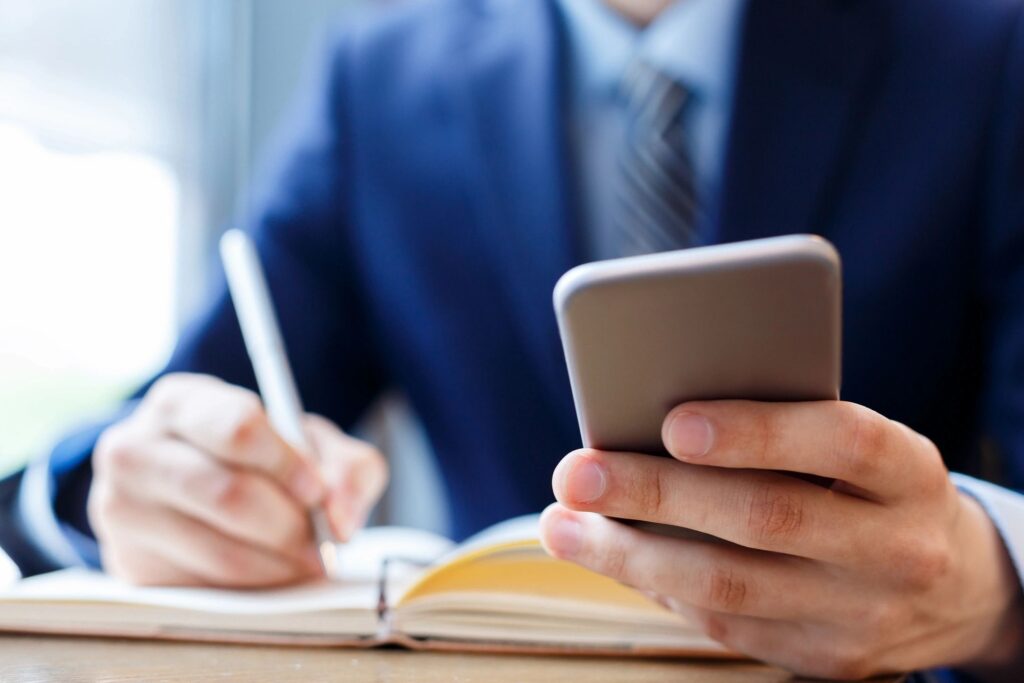 Businessman using smartphone while writing notes in a notebook.